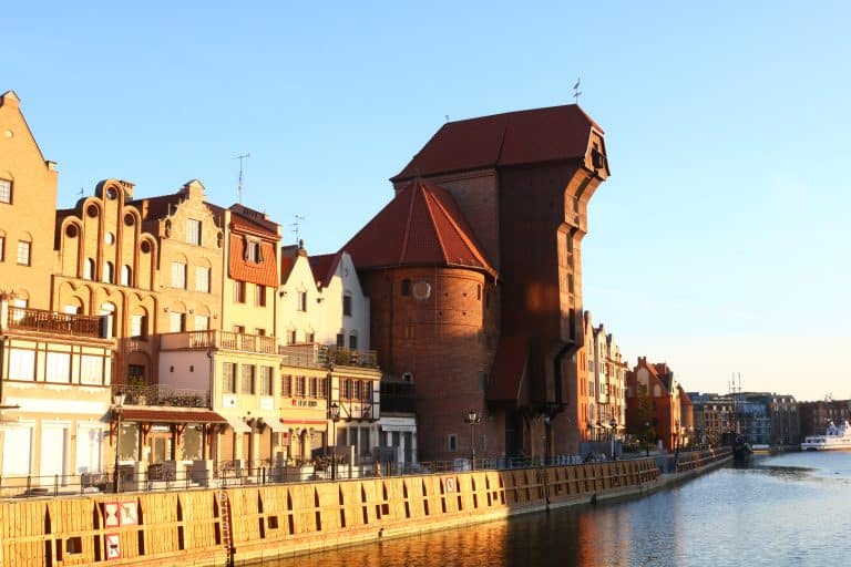 Historic Gdańsk waterfront at golden hour, featuring the medieval crane, pastel buildings, and reflections shimmering on the Motława River under a glowing sky.