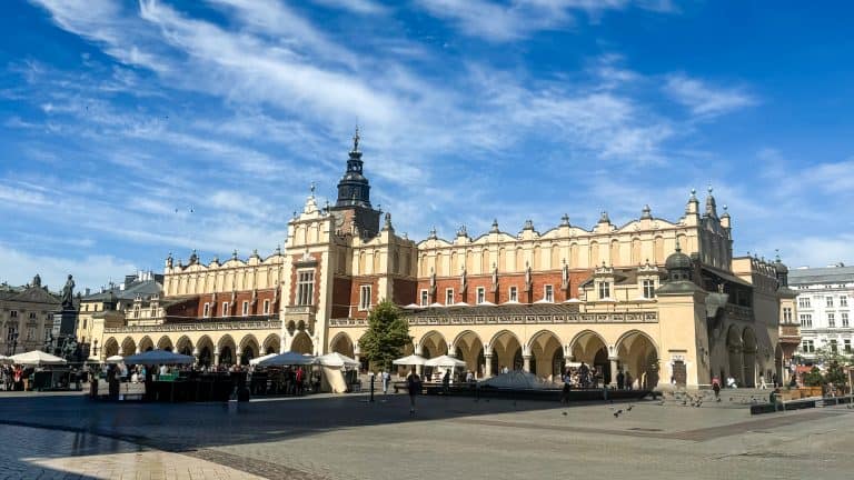 View of Kraków’s Main Square featuring the Cloth Hall, with arched arcades, a central tower, and people enjoying the open plaza under a partly cloudy sk