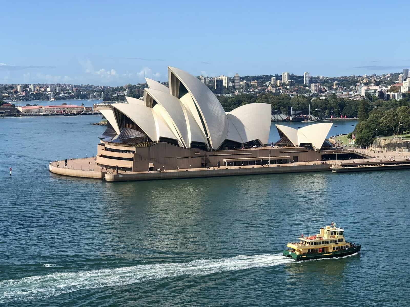 Sydney Opera House with its iconic white sail-like roofs viewed from across the harbor, with a ferry passing in the foreground under clear blue skies.
