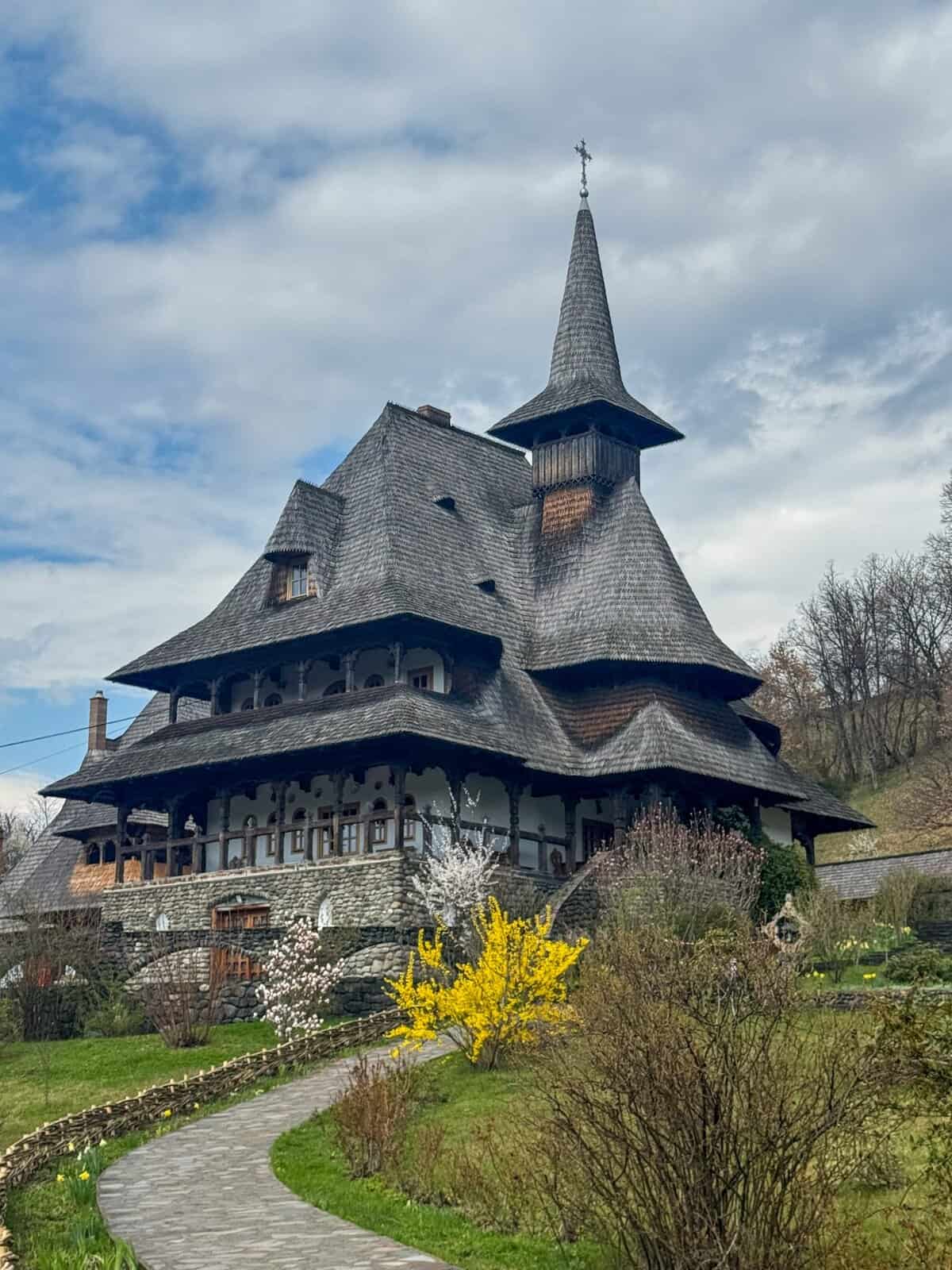 Traditional wooden church in Maramureș, Romania, with a tall steeple, steep roofs, and carved details, surrounded by flowers and a stone path