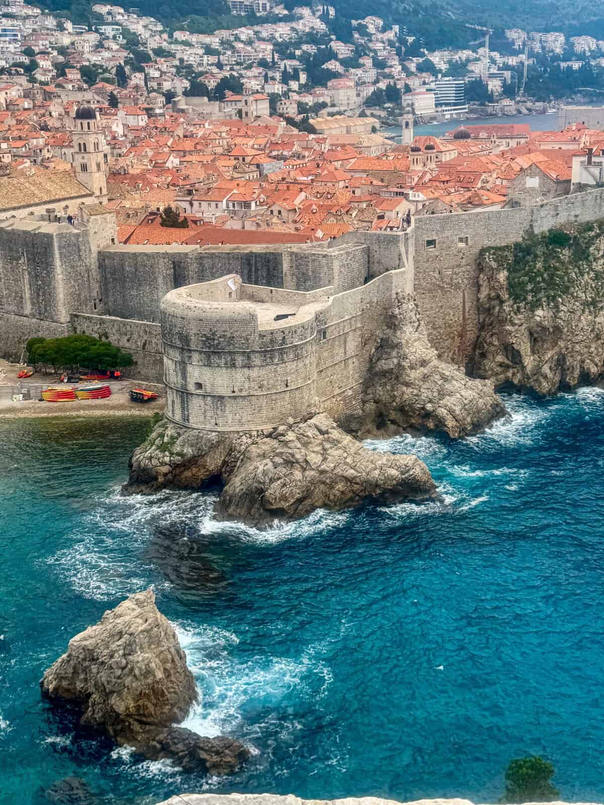 Panoramic view of Dubrovnik’s terracotta rooftops enclosed by medieval stone walls, with the Adriatic Sea shimmering in the foreground under a clear blue sky